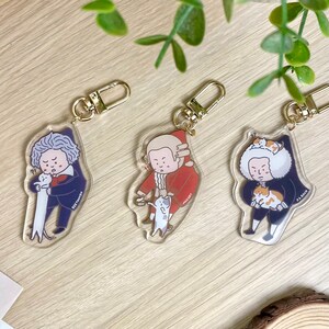 May include: Three clear acrylic keychains featuring cartoon illustrations of famous composers: Beethoven, Mozart, and Bach. Each composer is depicted holding a cat.