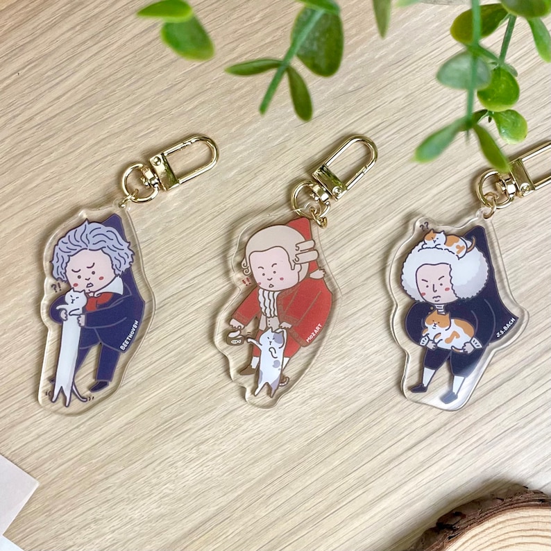 May include: Three clear acrylic keychains featuring cartoon illustrations of famous composers: Beethoven, Mozart, and Bach. Each composer is depicted holding a cat.