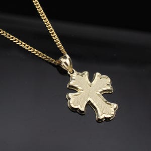 May include: A gold-colored pendant necklace with a cross-shaped charm. The charm has a textured surface and a detailed design. The chain is also gold-colored and has a curb link style. The necklace is displayed against a black background.