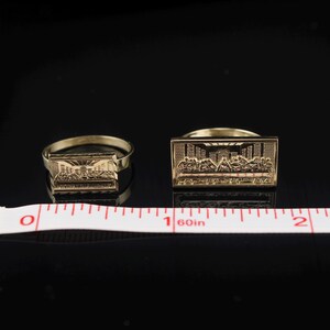 May include: Two gold-colored rings with rectangular faces, each depicting a detailed scene. The rings are placed on a black surface with a measuring tape. The rings are likely jewelry.