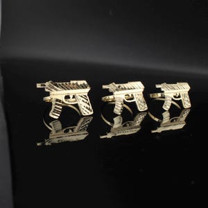 May include: Three gold-colored rings, each designed in the shape of a handgun. The rings are displayed on a reflective black surface, creating mirrored reflections of the jewelry. The rings have detailed textures and are evenly spaced.