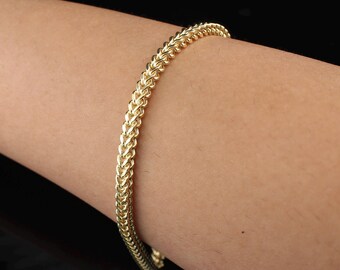 May include: A gold-colored, curb chain bracelet. The bracelet is made of interlocking links, creating a textured surface. The jewelry is displayed on a person's arm against a dark background. The bracelet is a classic piece of jewelry.