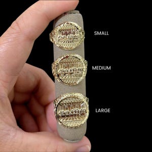 May include: Gold-colored rings in small, medium, and large sizes, each featuring a detailed relief of a religious scene. The rings are displayed on a gray ring holder against a black background. The word "SMALL", "MEDIUM", and "LARGE" are written in white.