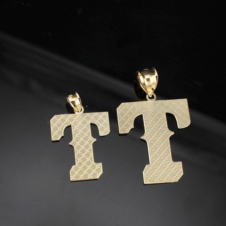 May include: Two gold-colored letter "T" pendants. The pendants are in different sizes, both with a textured surface. The pendants have a loop at the top for a chain. The background is black.
