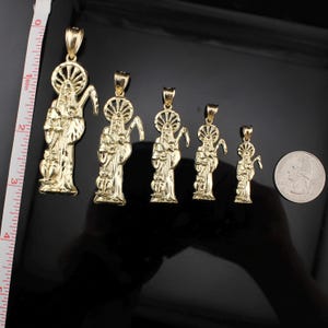 May include: A collection of gold-colored pendant necklaces in various sizes, featuring a religious figure with a halo and a scythe. The pendants are displayed on a reflective black surface, with a ruler showing inches for scale.
