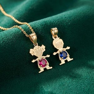 May include: Two gold-colored pendant necklaces. One pendant is a girl figure with a pink gemstone, the other is a boy figure with a blue gemstone. The pendants are on a gold chain, set against a dark green background.