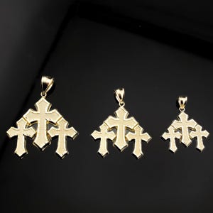 May include: Three gold-colored cross pendants in varying sizes, each featuring a cluster of three crosses. The pendants are displayed against a dark background, highlighting their intricate design and metallic sheen. The jewelry is suitable for religious or fashion purposes.