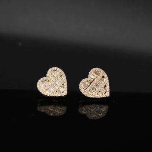 May include: A pair of heart-shaped stud earrings. The earrings are gold-toned and encrusted with small, sparkling, rectangular-cut gemstones. The gemstones are arranged in a diagonal pattern across the heart shape.