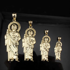 May include: Four gold-colored pendants featuring a religious figure with a halo. The pendants vary in size, with the largest on the left and the smallest on the right. Each pendant depicts a figure holding a staff and wearing draped robes.