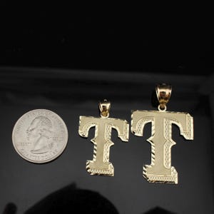 May include: Two gold-colored letter "T" pendants, one large and one small, with textured surfaces and bale attachments. A U.S. quarter coin is next to the pendants for size comparison. The pendants are displayed on a black surface.