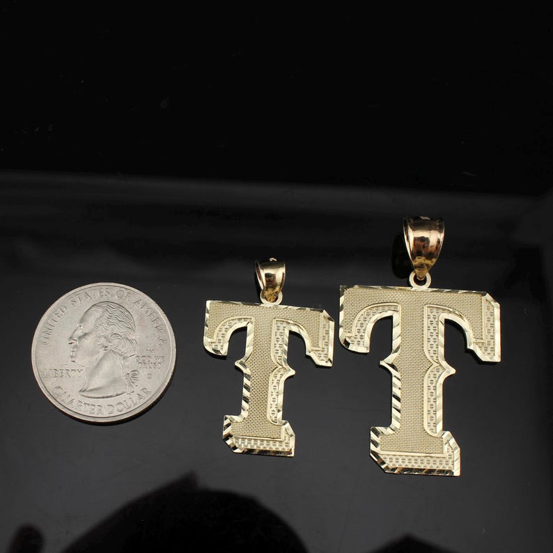 May include: Two gold-colored letter "T" pendants, one large and one small, with textured surfaces and bale attachments. A U.S. quarter coin is next to the pendants for size comparison. The pendants are displayed on a black surface.
