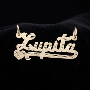 May include: A gold-colored pendant necklace with the name "Lupita" in a cursive font. The pendant has a heart-shaped design and is suspended from two small loops.