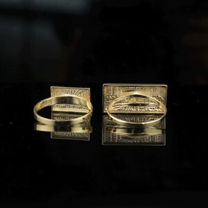 May include: Two gold-colored rings with rectangular faces. The rings are set against a black background, with their reflections visible. The rectangular faces have a detailed, textured design.