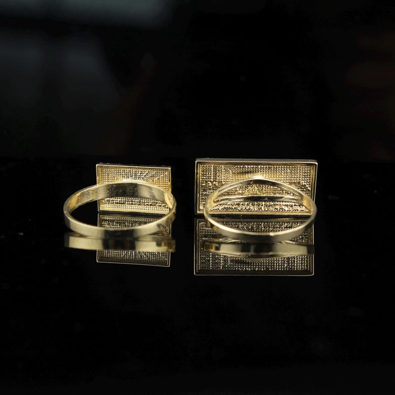 May include: Two gold-colored rings with rectangular faces. The rings are set against a black background, with their reflections visible. The rectangular faces have a detailed, textured design.