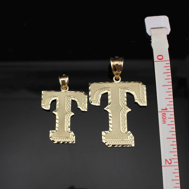 May include: Two gold-colored letter "T" pendants with textured surfaces and decorative edges. The pendants are displayed next to a measuring tape, with the larger pendant approximately 1.75 inches in height. The background is black.