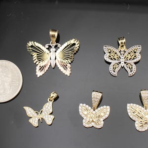 K&ouml;nnte beinhalten: F&uuml;nf goldfarbene Schmetterlingsanh&auml;nger mit unterschiedlichen Designs. Ein Anh&auml;nger hat ein dreifarbiges Fl&uuml;geldesign, andere sind mit klaren Edelsteinen verziert. Eine M&uuml;nze dient als Gr&ouml;&szlig;envergleich.