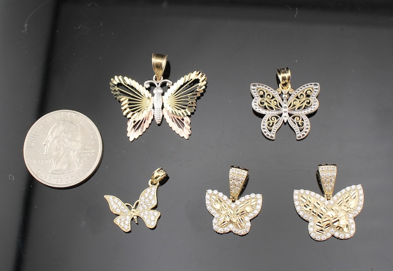 K&ouml;nnte beinhalten: F&uuml;nf goldfarbene Schmetterlingsanh&auml;nger mit unterschiedlichen Designs. Ein Anh&auml;nger hat ein dreifarbiges Fl&uuml;geldesign, andere sind mit klaren Edelsteinen verziert. Eine M&uuml;nze dient als Gr&ouml;&szlig;envergleich.