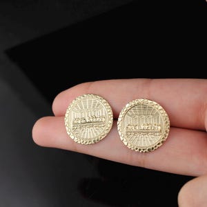 May include: Two gold-colored round pendants, each featuring a detailed relief of a religious scene. The pendants are held in a hand against a dark background. The design includes a detailed border.