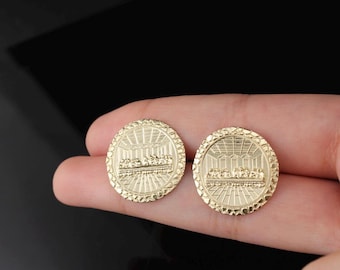 May include: Two gold-colored round pendants, each featuring a detailed relief of a religious scene. The pendants are held in a hand against a dark background. The design includes a detailed border.