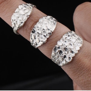 May include: Three silver-colored nugget rings are displayed on fingers against a dark background. The rings have a textured, irregular surface, resembling small, fused stones. The rings are a bright, shiny silver.