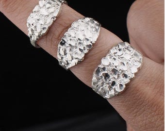 May include: Three silver-colored nugget rings are displayed on fingers against a dark background. The rings have a textured, irregular surface, resembling small, fused stones. The rings are a bright, shiny silver.