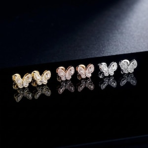May include: Six pairs of butterfly-shaped stud earrings. The earrings are arranged in a row and feature a variety of metal finishes, including gold, rose gold, and silver. Each earring is encrusted with small, sparkling gemstones, creating a shimmering effect.