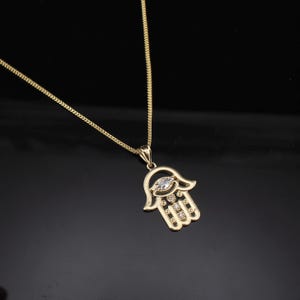 May include: Gold-toned necklace featuring a Hamsa hand pendant. The pendant has a central marquise-cut clear stone and smaller round clear stones. The chain is a delicate curb link style, and the background is black.