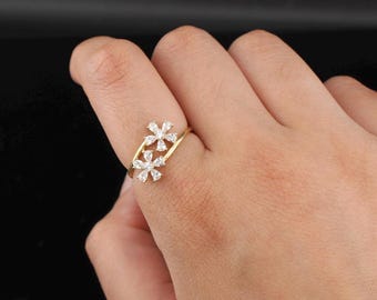 May include: A gold-colored ring featuring two flower-shaped designs. Each flower is composed of multiple small, clear gemstones. The ring is worn on a finger, with a black background.