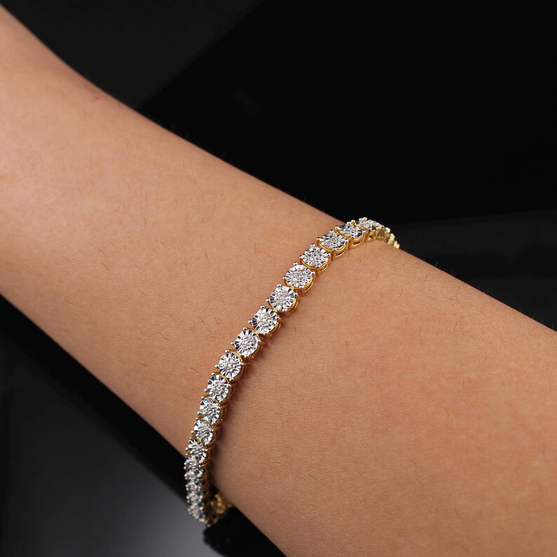 May include: A gold-toned bracelet with a series of round, clear gemstones. The bracelet is set on a gold band and is displayed on a person's arm against a black background. The gemstones are evenly spaced.