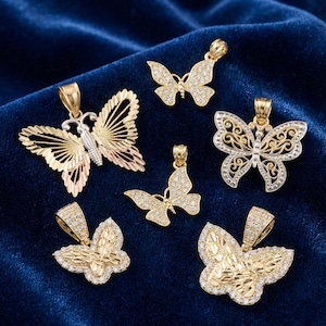 K&ouml;nnte beinhalten: Eine Sammlung goldener Schmetterlingsanh&auml;nger mit verschiedenen Designs, einige mit kleinen, funkelnden Steinen verziert. Die Anh&auml;nger werden auf einer dunkelblauen Samtoberfl&auml;che pr&auml;sentiert, die ihre komplizierten Details hervorhebt.