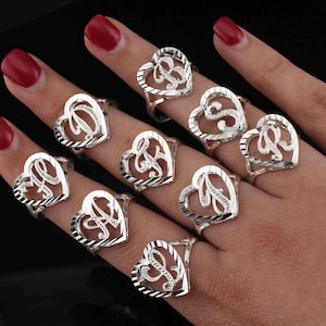 May include: A collection of silver-colored heart-shaped rings, each featuring a different initial letter. The rings are displayed on fingers against a dark background. The heart design has a textured, openwork pattern.