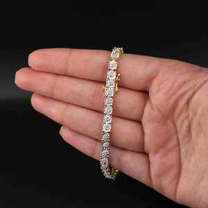 May include: A gold bracelet with a row of square-cut, clear gemstones. The bracelet is held in a hand against a black background. The clasp is visible, and the bracelet appears to be new.