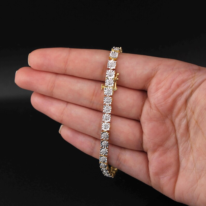 May include: A gold bracelet with a row of square-cut, clear gemstones. The bracelet is held in a hand against a black background. The clasp is visible, and the bracelet appears to be new.