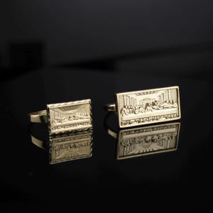 May include: Gold-toned cufflinks featuring a detailed relief of the Last Supper scene. The rectangular cufflinks have a polished finish and a classic design, suitable for formal wear.