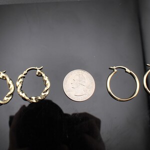 May include: Two pairs of gold-colored hoop earrings. One pair features a twisted rope design, while the other pair has a smooth, classic hoop style. A US quarter is placed between the earrings for size comparison.