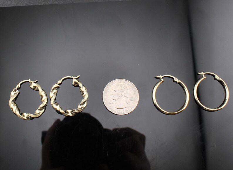 May include: Two pairs of gold-colored hoop earrings. One pair features a twisted rope design, while the other pair has a smooth, classic hoop style. A US quarter is placed between the earrings for size comparison.