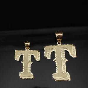 May include: Two gold-colored letter "T" pendants. The pendants are in a serif font with a textured surface. The pendants have a loop at the top for a chain. The background is black.
