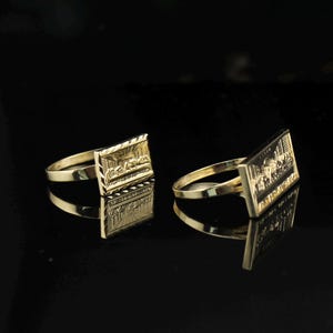 May include: Two gold-colored rings with rectangular faces, each featuring a detailed relief design. The rings are set against a black background, with reflections visible. The rings appear to be made of metal.