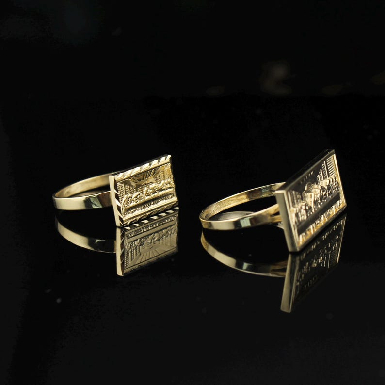May include: Two gold-colored rings with rectangular faces, each featuring a detailed relief design. The rings are set against a black background, with reflections visible. The rings appear to be made of metal.