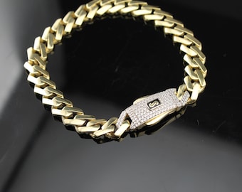 May include: A gold-colored chain bracelet with a diamond-studded clasp. The bracelet features a thick, curb link design. The clasp has a small, black and white emblem. The bracelet is displayed on a reflective black surface.