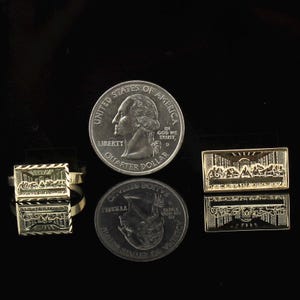 May include: A gold-colored ring and a rectangular pin featuring a detailed relief of a religious scene. A U.S. quarter coin is next to the ring and pin for size comparison. The ring and pin are on a reflective black surface.