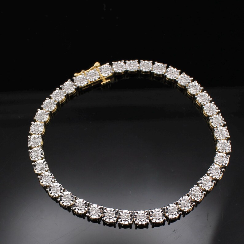 May include: A gold-toned tennis bracelet with a series of round, sparkling diamond-like stones. The bracelet is set against a dark background, highlighting the brilliance of the stones and the gold clasp.