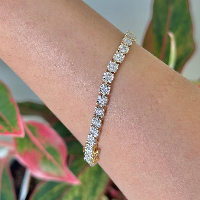 May include: A gold tennis bracelet featuring a series of round, clear gemstones. The bracelet is clasped on a wrist, showcasing the sparkle of the stones. The background includes green and pink foliage.