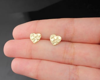 May include: A pair of gold-colored heart-shaped stud earrings. The earrings have a textured surface and are displayed on a light-toned background. The earrings are small and delicate, perfect for everyday wear.