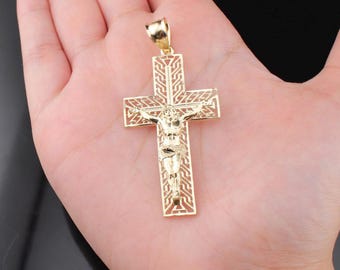 May include: Gold-colored pendant in the shape of a cross, featuring a detailed depiction of Jesus Christ. The cross has an openwork design with intricate patterns. The pendant is held in a person's hand against a dark background.