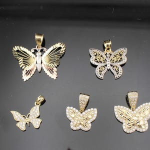 K&ouml;nnte beinhalten: F&uuml;nf Schmetterlingsanh&auml;nger in Gold- und Silbert&ouml;nen. Die Anh&auml;nger weisen verschiedene Designs auf, darunter offene Fl&uuml;gel, filigrane Muster, und einige sind mit kleinen, funkelnden Steinen verziert. Die Schmetterlinge werden vor einem dunklen Hintergrund pr&auml;sentiert.