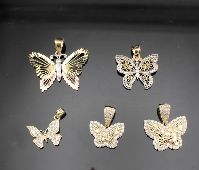 K&ouml;nnte beinhalten: F&uuml;nf Schmetterlingsanh&auml;nger in Gold- und Silbert&ouml;nen. Die Anh&auml;nger weisen verschiedene Designs auf, darunter offene Fl&uuml;gel, filigrane Muster, und einige sind mit kleinen, funkelnden Steinen verziert. Die Schmetterlinge werden vor einem dunklen Hintergrund pr&auml;sentiert.