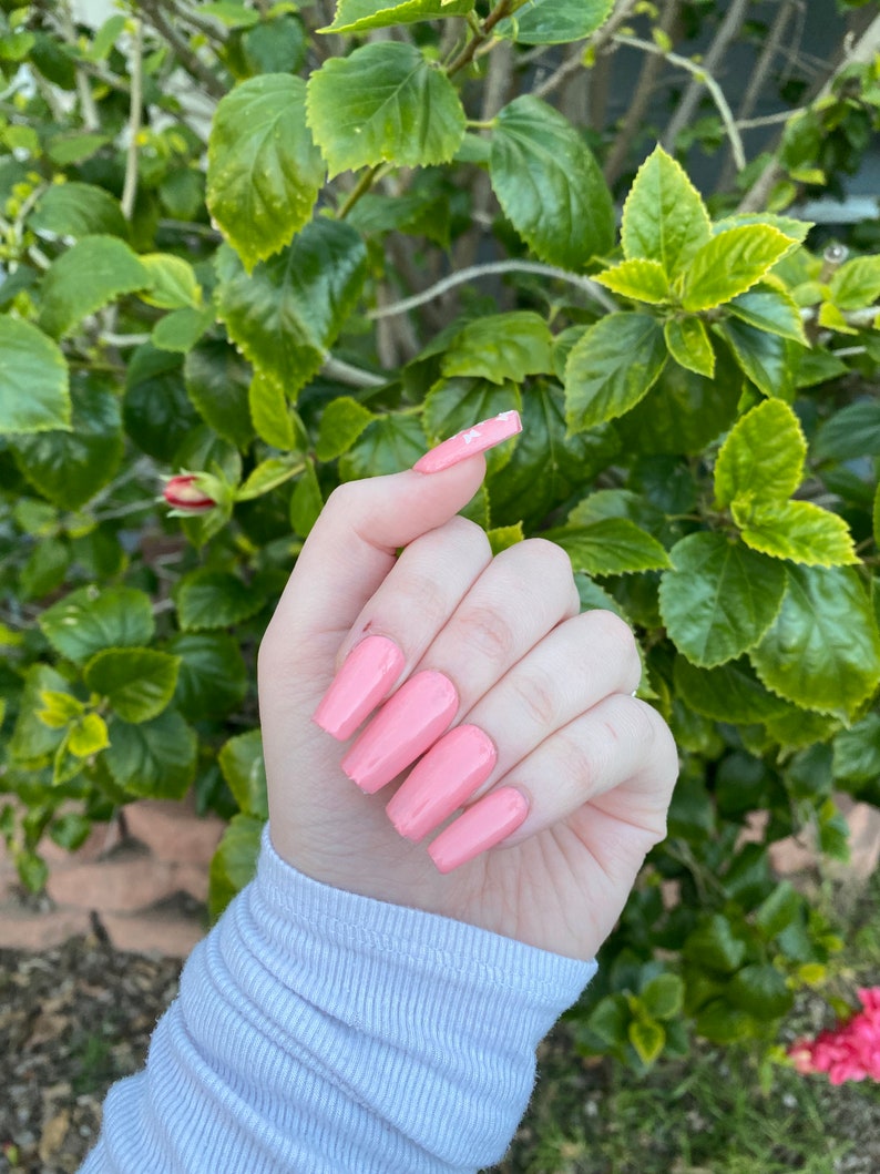 Op de afbeelding: Close-up van een hand met lange, roze, amandelvormige nagels. De nagels zijn geverfd in een lichtroze kleur en hebben een glanzende afwerking.