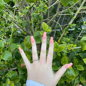 Op de afbeelding: Een hand met lange, vierkante, lichtroze nagels met een kleine, heldere, glinsterende ring aan de ringvinger. De nagels hebben een subtiele glans.