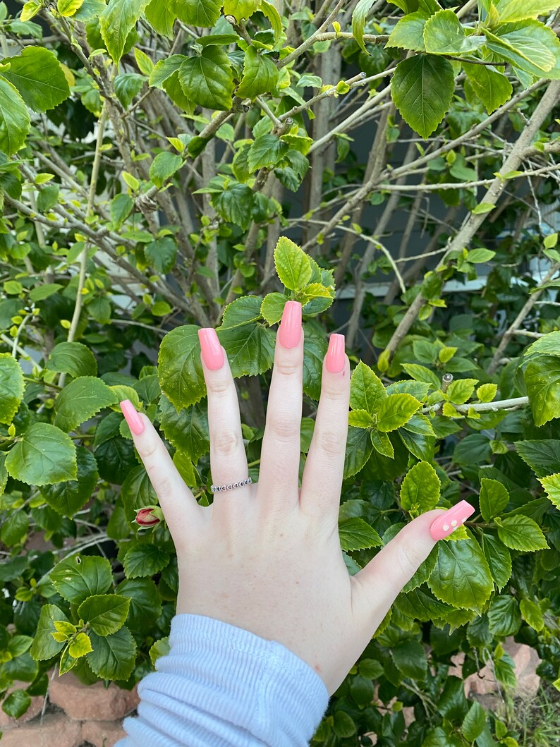 Op de afbeelding: Een hand met lange, vierkante, lichtroze nagels met een kleine, heldere, glinsterende ring aan de ringvinger. De nagels hebben een subtiele glans.